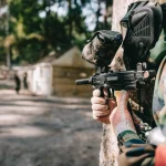 selective focus of paintball player in goggle mask and camouflage aiming by paintball gun outdoors
