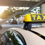 Close-up detail yellow taxi symbol on cars roof stand waiting at parking of airport terminal or railway station against park warm evening bokeh sunlight. Urban street transportation comfort service